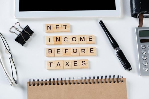 Net income before taxes concept with letters on cubes. Still life of office workplace with supplies. Flat lay white surface with tablet computer and calculator. Financial management and taxation.