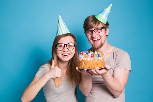 Funny young couple in paper caps and with a cake make a foolish face and wish happy birthday while standing against a blue background. Concept of congratulations and fooling around