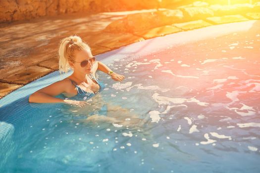 Woman in blue swimsuit and sunglasses relaxing in outdoor pool with clean transparent turquoise water. Woman sunbathing in bikini at tropical resort. Sun flare