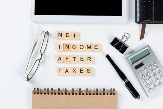 Net income after taxes concept with letters on cubes. Still life of office workplace with supplies. Flat lay white surface with tablet computer and calculator. Financial management and taxation.