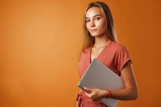 Serious young blonde woman standing with laptop and looking at camera against orange background