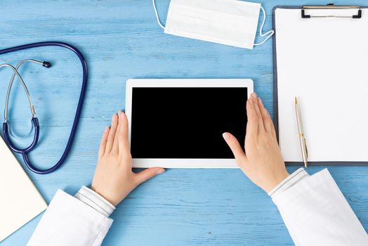 Top view of doctor hands with tablet computer. Therapist sitting at blue desk with stethoscope and clipboard. Examination and consultation in clinic. Medical application and online healthcare services