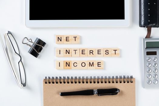 Net interest income concept with letters on wooden cubes. Still life of office workplace with supplies. Flat lay white surface with tablet computer and notepad. Financial management and accounting