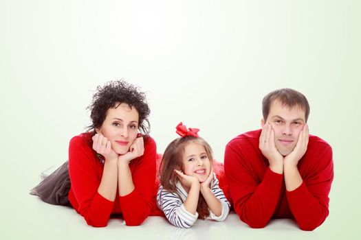 Happy young family dad mom and a little girl in bright red outfits . Family lying on the floor leaning on his hands.Summer white green blurred background.