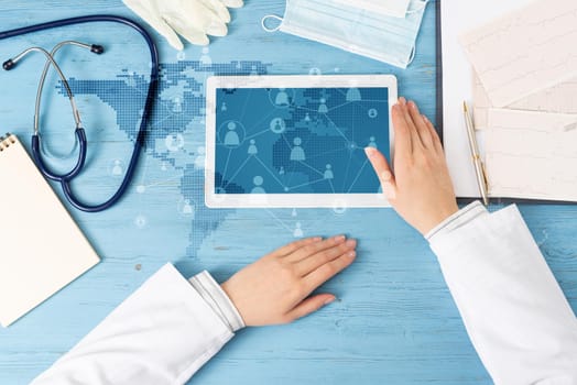 Top view of doctor hands with tablet computer. Therapist in white coat sitting at blue wooden desk. Examination and consultation in clinic. Medical application and online healthcare services.