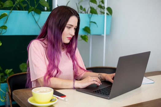 Young woman with pink hair with laptop computer sitting in cafe, intelligent female student working on net-book after her lectures in University