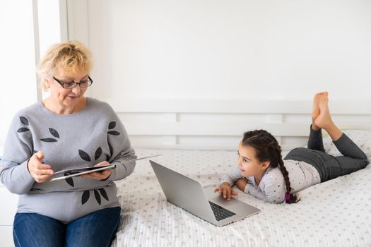 Mature grandmother helping child with homework at home. Satisfied old grandma helping her granddaughter studying in living room. Little girl writing on notebook with senior teacher sitting next to her