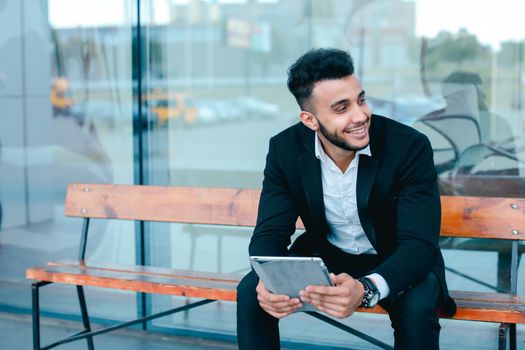 Entrepreneur smiling man puts on sunglasses uses tablet and looks at. Young handsome businessman arab muslims in business center wearing dressed in black elegant suit on building background.