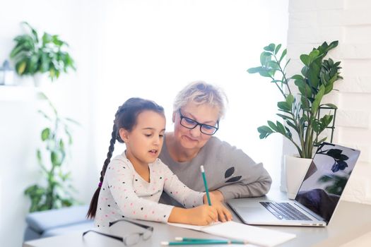 Mature grandmother helping child with homework at home. Satisfied old grandma helping her granddaughter studying in living room. Little girl writing on notebook with senior teacher sitting next to her