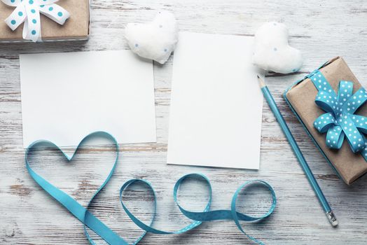 Flat lay composition with blue ribbon in shape of heart. Blank sheet of white paper and gift box on vintage wooden table. Happy birthday or anniversary congratulation. Romantic event invitation.