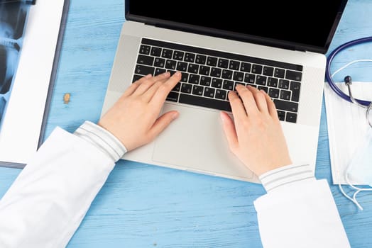 Top view of doctor hands typing at laptop computer. Therapist sitting at blue wooden desk with x-ray scan. Examination and consultation in clinic. Medical application and online healthcare services.