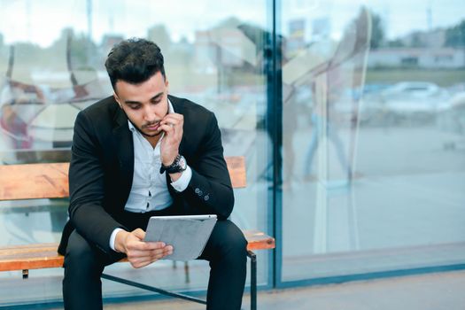 Entrepreneur smiling man puts on sunglasses uses tablet and looks at. Young handsome businessman arab muslims in business center wearing dressed in black elegant suit on building background.