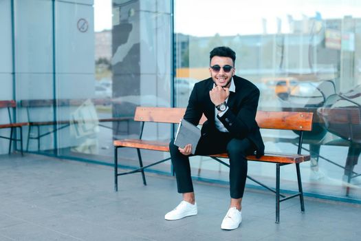 Entrepreneur smiling man puts on sunglasses uses tablet and looks at. Young handsome businessman arab muslims in business center wearing dressed in black elegant suit on building background.