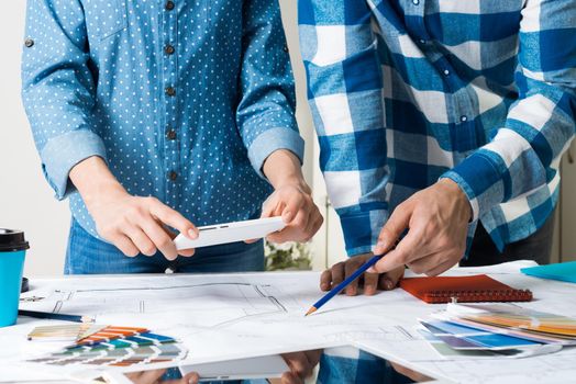 Woman taking photo of technical drawing with smartphone. Designers working with color swatches and construction blueprint at workplace. Interior design, architectural decoration and renovation.