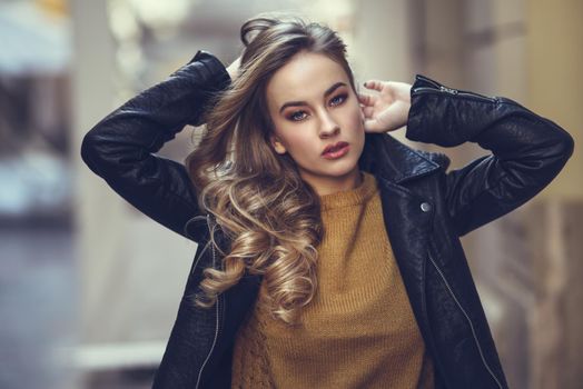 Blonde woman in urban background. Beautiful young girl wearing black leather jacket and mini skirt standing in the street. Pretty russian female with long wavy hair hairstyle and blue eyes.