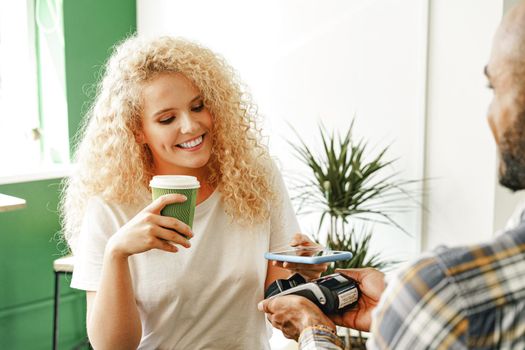 Woman customer of coffee shop paying for coffee through mobile phone using contactless technology close up