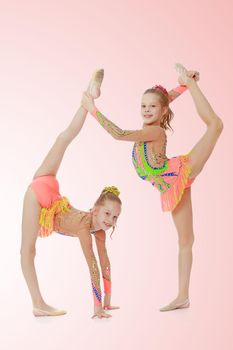 Two adorable little twin girls, gymnastics in the sports school. Girls beautiful gymnastic leotards. They do the splits.Pale pink gradient background.