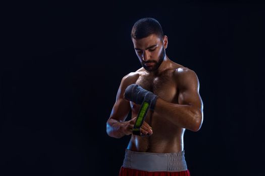 Sports boxer man pulls on the hand wrist wraps. Oriental male model isolated on black background. Athletic man posing in red shorts without shirt