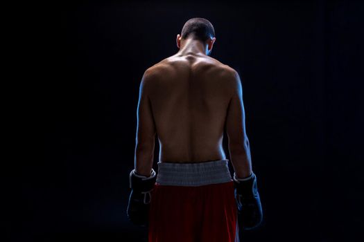 A boxer in his shorts stands with his back, his head is lowered. Recreation. Black background. Strength and motivation. Studio shot
