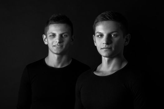 Two attractive positive young twin brothers standing dressed in black sweaters on black background. Black and white photography