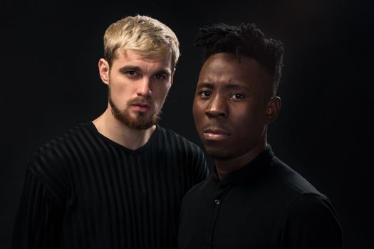 Portrait of two young african american and caucasian men standing over black background. International friendship concept. Studio shot