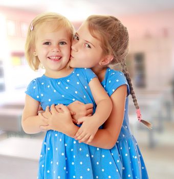 Two charming little girls, sisters , in identical blue dresses with polka dots. Older sister kissing the younger on the cheek.On the background of the room where children live, and are on the shelves of toys.