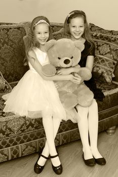 Two adorable little twin girls sitting on sofa hugging a big Teddy bear.Black-and-white photo. Retro style.