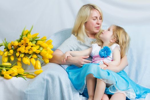 Mother and daughter in identical blue dresses hugging sitting on the sofa.Next to them lies a large bouquet of yellow tulips.