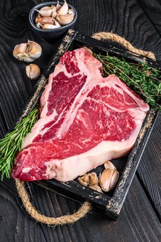 Porterhouse or T bone raw beef meat Steak with herbs and salt in wooden tray. Black wooden background. Top view.