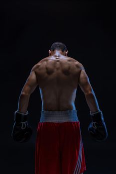 Rear view of strong young male boxer. Fitness male model wearing boxing gloves standing on black background. Studio shot
