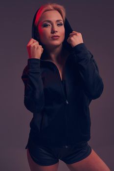Young pretty woman wearing a black hood and shorts on dark background. olor filters. Studio shot. A woman looks at the camera