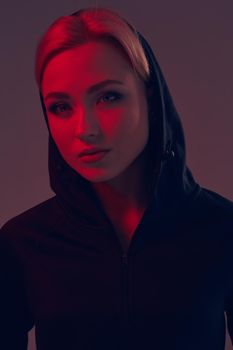 Young pretty woman wearing a black hood and shorts on dark background. olor filters. Studio shot. A woman looks at the camera. Portrait