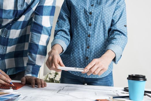 Woman taking photo of technical drawing with smartphone. Designers working with color swatches and construction blueprint at workplace. Interior design, architectural decoration and renovation.