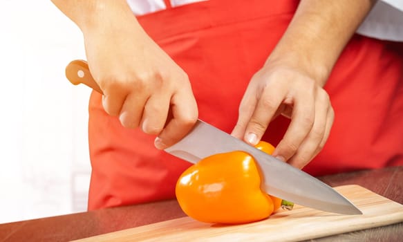 Chef hands cutting orange fresh pepper on wooden board. Close up man hands cooking with knife on table in kitchen. Professional chef in red apron on light background. Restaurant food preparation.