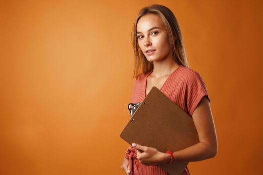 Young beautiful smiling woman standing and holding clipboard against orange background