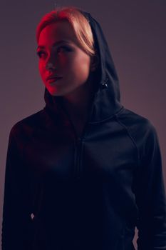Young pretty woman wearing a black hood and shorts on dark background. olor filters. Studio shot.