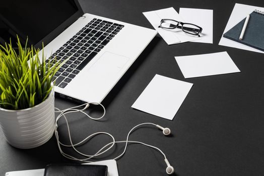 Top view of office workplace with digital gadgets. Flat lay black surface with laptop, green plant and white paper sheets. Top view coworking workspace with devices. Digital technology and mobility.