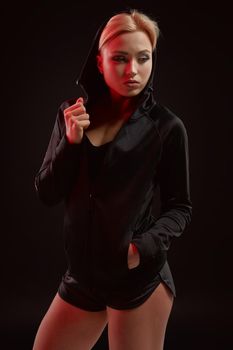 Beautiful seductive sporty girl with the slim sexy figure in the black hoodie, sport underwear and little black shorts is posing in the studio. olor filters.