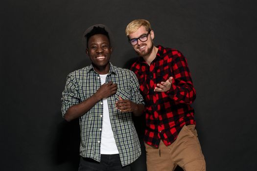 Studio shot of two stylish young men having fun. Handsome bearded hipster in a shirt in a cage standing next to his African-American friend in hat against a dark background. International friendship concept.