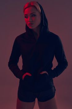 Beautiful seductive sporty girl with the slim sexy figure in the black hoodie, sport underwear and little black shorts is posing in the studio. color filters.