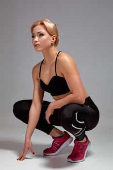 Happy cute young sportswoman sitting over gray background. Studio shot. Young woman wears black leggings and sports top