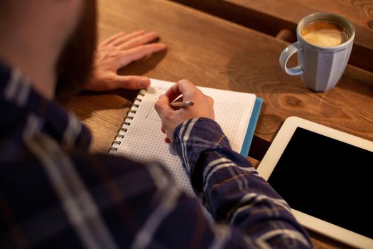Young bearded businessman sits in cafe, home at table and writes in notebook, near lies tablet computer with black screen. Man is working, studying. Online education, marketing, training.