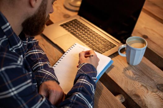Cropped image of casual business man or freelancer planning his work on notebook, working on laptop computer with smart phone, cup of coffee on table at coffee shop or home office, working from cafe concept