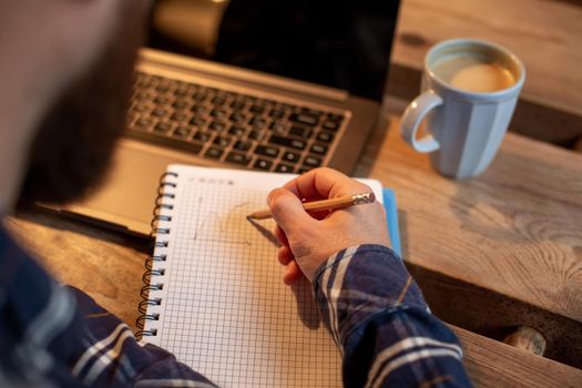 Cropped image of casual business man or freelancer planning his work on notebook, working on laptop computer with smart phone, cup of coffee on table at coffee shop or home office, working from cafe concept