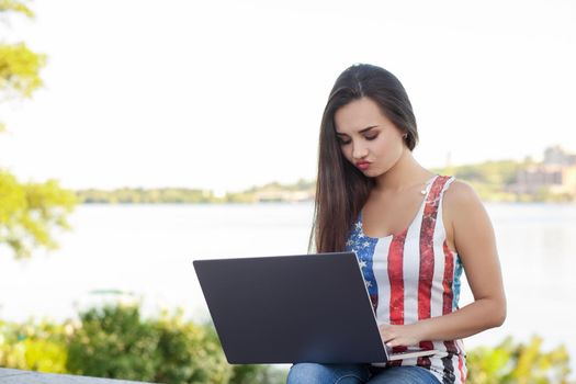 The beautiful young woman sits in the park near river with the laptop. Success small business, modern lifestyle, information technology, or online shopping concept