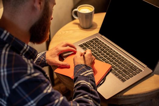 Cropped image of casual business man or freelancer planning his work on notebook, working on laptop computer with smart phone, cup of coffee on table at coffee shop or home office, working from cafe concept