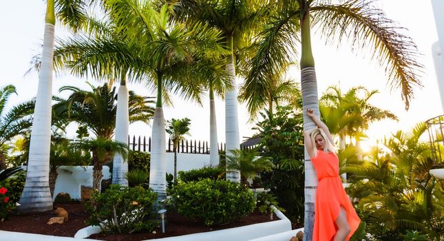 Close-up portrait of joyful laughing young lady in orange dress posing with hand up near the open-air, on palm trees background