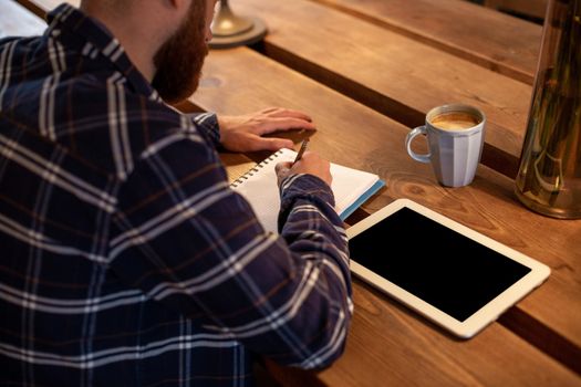 Young bearded businessman sits in cafe, home at table and writes in notebook, near lies tablet computer with black screen. Man is working, studying. Online education, marketing, training.