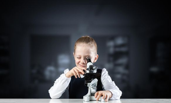 Little girl scientist looking through microscope in classroom at biology lesson. Research and discovery. Formal education in modern school. Schoolgirl in schoolwear making experiment in class.