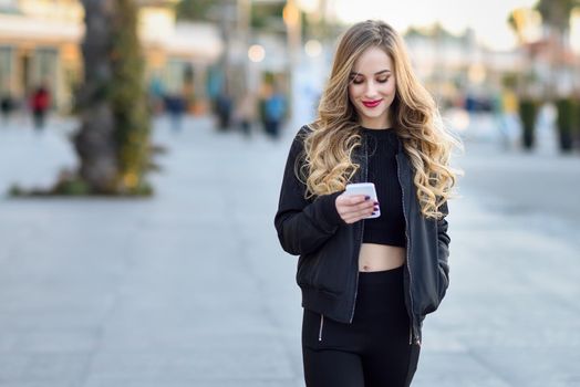 Blonde woman texting with her smart phone in urban background. Beautiful young smiling girl wearing black jacket walking in the street. Pretty russian female with long wavy hair hairstyle.